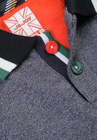 Grijze polo met een gestructureerde afwerking, zwarte kraag, rode en groene knopen, en een levendige rode binnenlabel met het merk J&Joy.