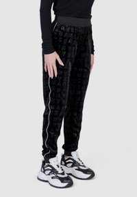 Pantaloni joggers in velluto nero con stampa del logo texture, caratterizzati da strisce laterali bianche. Abbinati a sneakers chunky in bianco e nero.