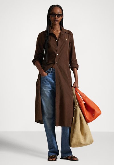 Woman with long braids wearing sunglasses, a brown long buttoned shirt, blue jeans, dark sandals, holding beige and orange tote bags.