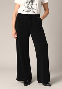 Zwarte wijde broek gemaakt van een zachte, textuurstof. Voorzien van een elastische tailleband met een trekkkoord en zijzakken.