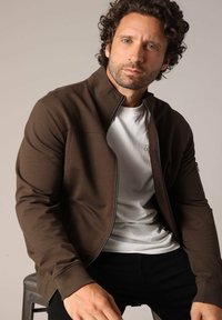 Veste zippée marron avec un col haut, fabriquée en tissu lisse. Portée sur un t-shirt blanc, avec un détail de poche zippée sur le côté.