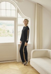 Black athletic set featuring a long-sleeve top with beige side stripes and matching pants. Worn with gray and beige sneakers. Natural wood floor.