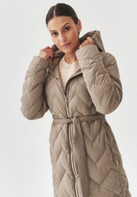 Beige vuorattu puffatakki, jossa on huppu, vetoketjukiinnitys ja vyötäröllä solmittava vyö. Ominaisuuksiin kuuluu timanttikuvio ja ribatty vyö lisäyksinä.