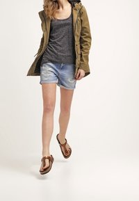 Olivově zelená parka, šedý tílko, světlé modré dírkované kraťasy, hnědé žabky, model chůze, neutrální pozadí.