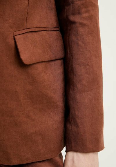 Close-up af brun linnedblazer-ærme og klaplomme på venstre side mod en neutral baggrund.