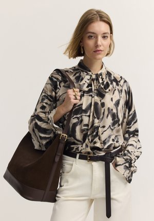 Frau trägt eine gemusterte Bluse, weiße Hose und goldene Ringe, trägt eine große braune Lederschultertasche vor hellem Hintergrund.