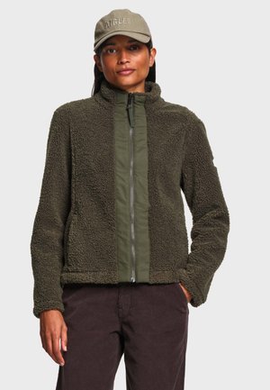 Femme portant une veste en polaire vert olive avec une fermeture éclair devant et une casquette vert olive, debout avec une main dans la poche d’un pantalon marron.