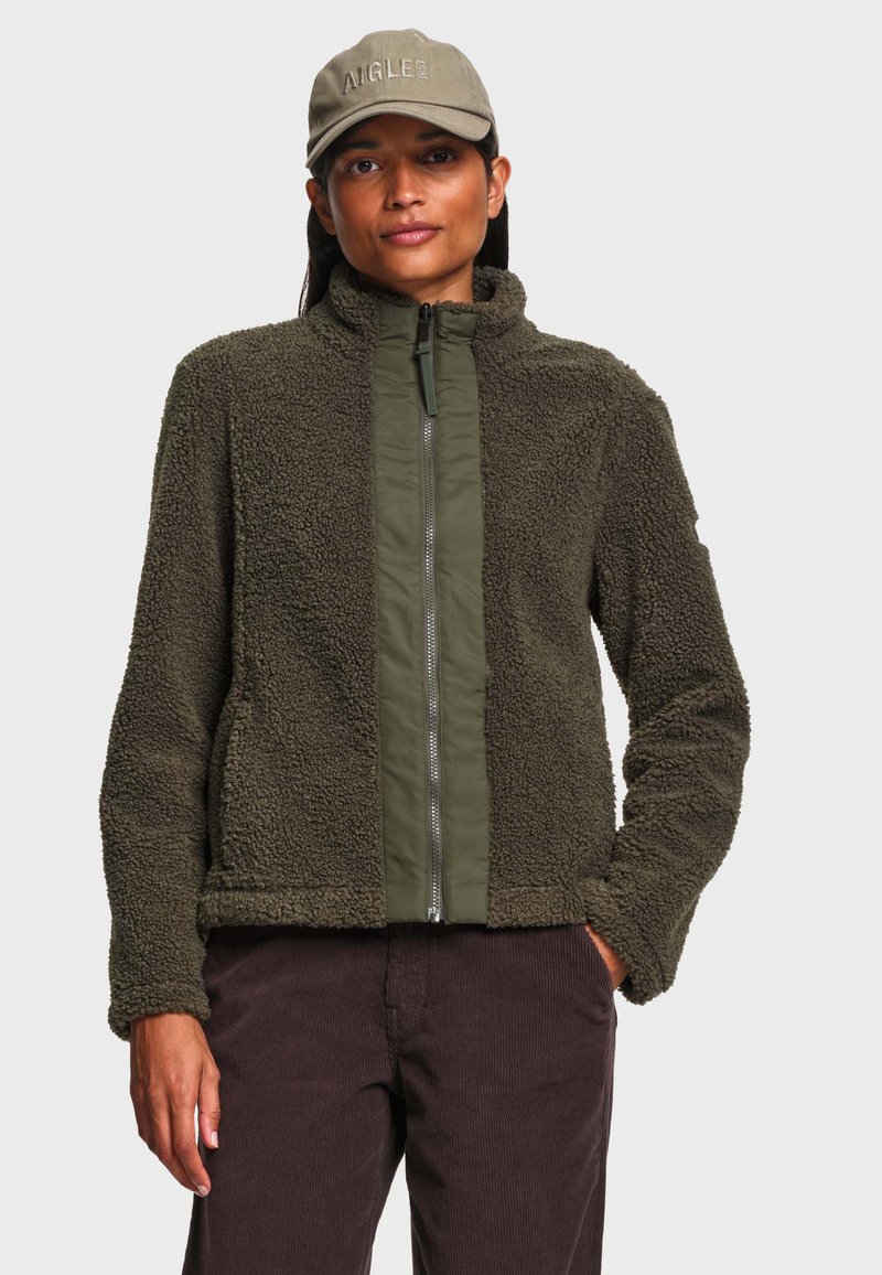 Femme portant une veste en polaire vert olive avec une fermeture éclair devant et une casquette vert olive, debout avec une main dans la poche d’un pantalon marron.