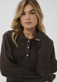 Tumši brūns ribots džempera ar apkakli, kurā ir trīs pērļu pogu aizdari un smalkas mirdzošas akcenti visā audumā.