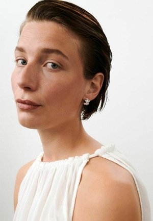 Nő rövid, nedves hajjal, fehér ujjatlan felsőt és apró virágmintás ezüst fülbevalókat visel, enyhén a vállai felett néz.