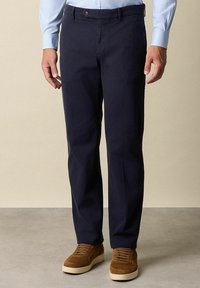 Muž v tmavě modrých rovně střižených kalhotách a hnědých semišových šněrovacích teniskách, stojící na šedé podlaze proti béžové zdi.