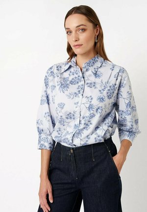 Frau mit glattem braunem Haar trägt ein blau-weiß geblümtes Hemd zum Knöpfen und dunkle hoch taillierte Jeans, steht mit einer Hand in der Tasche.