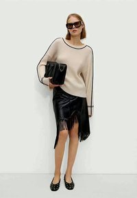Donna con maglione a coste beige, gonna in pelle nera con frange, ballerine nere con borchie, occhiali da sole neri di grandi dimensioni, mentre tiene una borsa nera contro un muro bianco.