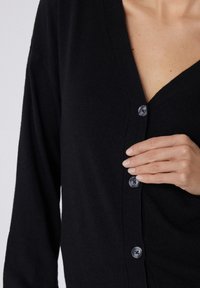 Schwarze Strickjacke mit V-Ausschnitt, geripptem Design und fünf Knopfverschlüssen. Der Stoff wirkt weich und hat eine figurbetonte Silhouette.