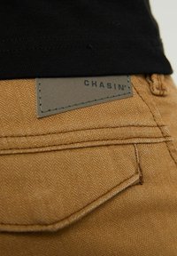 Tan herringbone-mönstrad tyg med en läderpatch märkt "CHASIN'". Närbild av sömmarna och bakfickans detaljer, utan synlig hårdvara.