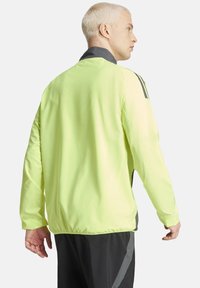 Veste de sport légère, vert fluo avec des accents noirs, à manches longues, ourlet élastique et col montant, fabriquée en matériau lisse et respirant.