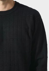 Maglione nero lavorato a maglia con un motivo a griglia, caratterizzato da un collo rotondo e texture a coste. Il tessuto sembra morbido e di peso medio.
