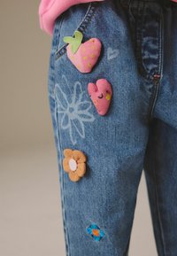 Enfant portant un jean bleu décoré de patchs roses en forme de fraise et de cœur, ainsi que des patchs fleurs orange et bleue sur la cuisse.