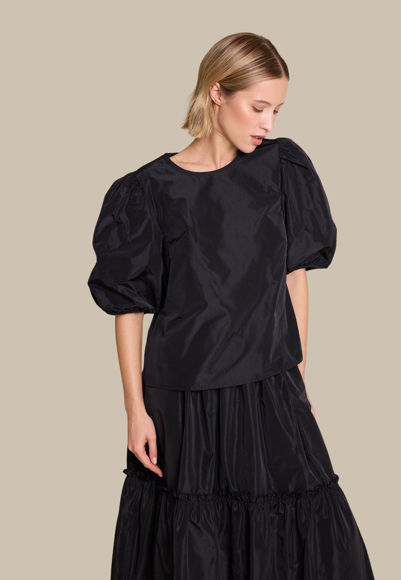 Svart topp med puffermer laget av glatt stoff, med rund halsutskjæring og avslappet passform. Kombinert med et lagdelt svart skjørt.