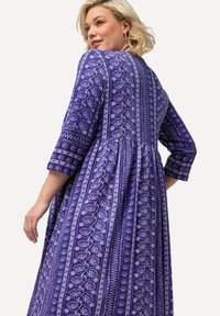 Violet mekko, jossa on kukka- ja geometrisia valkoisia kuvioita, kolmanneksen pituiset hihat ja levenevä hame kerättyine vyötärölinjoineen.