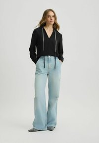 Zwarte langemouwtop met V-hals en zilveren kraaltjes als trekkoorden, gecombineerd met lichtblauwe wijde jeans met versleten details en patroon schoenen.