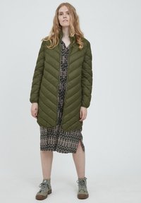 Olivenfarvet quilted puffer jakke med lynlås fortil, parret med en mønstret mørk kjole og grå ankelstøvler. Bemærkelsesværdige sidesnit i kjolen.