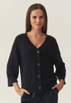 Mujer de cabello castaño liso, con un cárdigan negro de botones con cuello en V y mangas tres cuartos, con las manos en los bolsillos de unos pantalones oscuros.