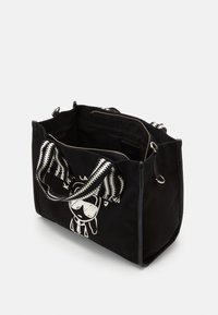 Černá taška přes rameno s bílým potiskem karikaturní tváře, pruhovanými textilními popruhy a vrchním zipem, který odhaluje vnitřní kapsy.