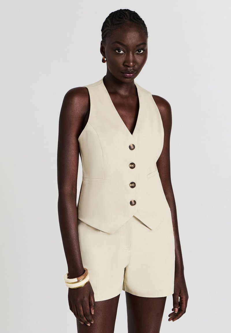 Frau mit kurzen geflochtenen Haaren, die eine beige ärmellose Weste mit Knöpfen und passende Shorts trägt, accessorized mit Armbändern und Ohrringen.