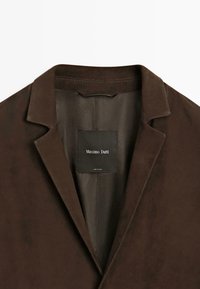 Bruine suède blazer met een notch-kraag, gladde voering en een label met de tekst "Massimo Dutti." Eenvoudig ontwerp met een getailleerde pasvorm.