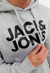 Nahaufnahme einer Person, die die Kordel des hellgrauen Hoodies mit großen schwarzen "JACK & JONES"-Schriftzug auf der Vorderseite anpasst.