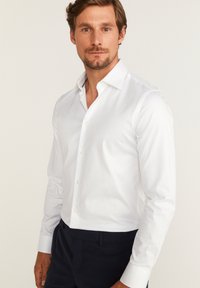 Witte langslavende overhemdd met een klassieke boord, knoopsluiting aan de voorkant en een slanke pasvorm. De stof lijkt soepel en heeft een lichte glans.