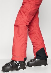Pantalons de ski isolés rouges avec un accent noir à la cheville et le logo Columbia. Associés à des bottes de ski noires avec des lettrages blancs et des boucles argentées.
