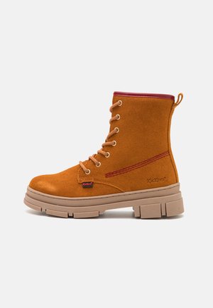 Bruine suède veterlaars met dikke beige zool en rode bies, zijaanzicht op effen achtergrond.