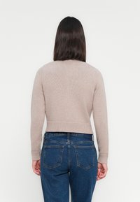 Vrouw met schouderlang donker haar, gekleed in een beige gebreide trui en blauwe jeans, met de rug naar de camera gekeerd tegen een effen witte achtergrond.
