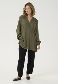 Haut à manches longues vert olive avec un col en V et un détail de bouton, en tissu texturé et avec des manches bouffantes, associé à un pantalon noir et des ballerines.