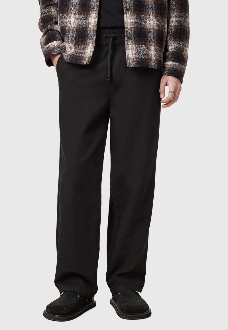 Pantalons noirs tissés avec une coupe décontractée, dotés d'une taille à cordon de serrage et de poches latérales, associés à une chemise à carreaux et des sandales noires.