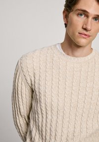 Beige Kabelstrickpullover mit rundem Halsausschnitt, ausgestattet mit strukturiertem Rippmuster und einem darüber liegenden weißen Hemd. Weiches Strickmaterial.