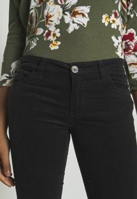 Kvinna i svarta kordbyxor och en grön blommig långärmad topp, som visar midjan och har handen vila vid sidan.