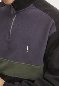 Sweatshirt de felpa con media cremallera que presenta paneles en morado oscuro y negro, una franja horizontal verde y un detalle de signo de exclamación en blanco que contrasta.