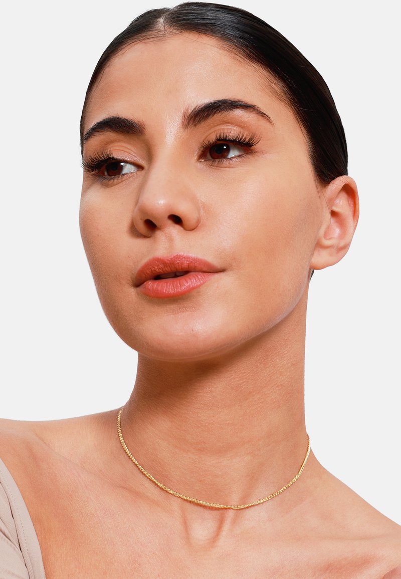 Collana choker delicata in tono oro con design intrecciato, poggiata su pelle nuda, che mette in risalto la sua texture liscia e il suo lieve splendore.