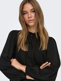 Svart blus med texturerat tyg, långa puffärmar, rund halsringning och knappdetalj framtill. Diskreta spetsdetaljer längs knappslån.