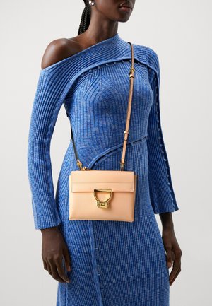 Donna che indossa un vestito blu a costine con una spalla scoperta e porta una piccola borsa a tracolla in pelle marrone chiaro con finiture dorate.