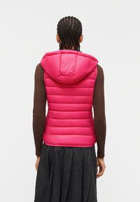 Άτομο με πλεξούδες φορά φωτεινό ροζ επενδεδυμένο μπουφάν με κουκούλα πάνω από καφέ μακρυμάνικο μπλουζάκι και σκούρα πλισέ φούστα, εμφανίζεται από πίσω.