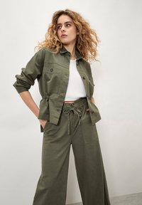 Chaqueta de algodón verde oliva con dos bolsillos en el pecho, cierre de botones y cintura elástica, combinada con pantalones de pierna ancha a juego con cordón.