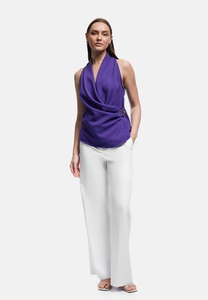 Kvinde iført en ærmeløs, lilla draperet bluse med sidelås og hvide bukser med vide ben, står med den ene hånd i lommen.