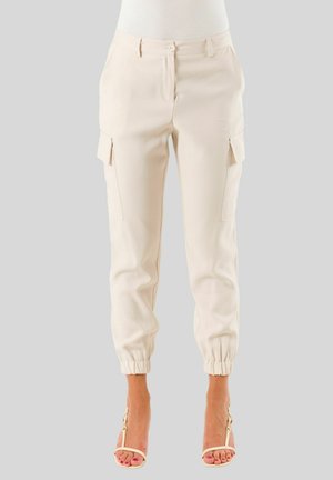 Pantaloni cargo color crema con tasche laterali e polsini elasticizzati, indossati con sandali beige con tacco alto su sfondo grigio.