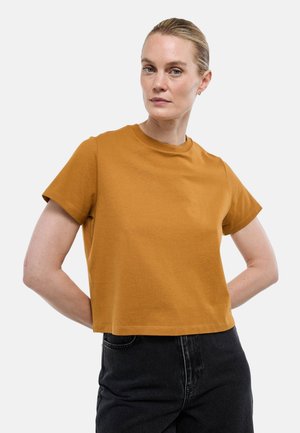 Femme aux cheveux blonds attachés, portant un t-shirt manches courtes moutarde et un pantalon noir taille haute, debout les mains derrière le dos.