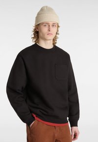 Sweatshirt noir avec un col rond, des poignets côtelés et une poche sur la poitrine gauche. Portez-le sur une chemise de couche rouge, associé à un pantalon marron.