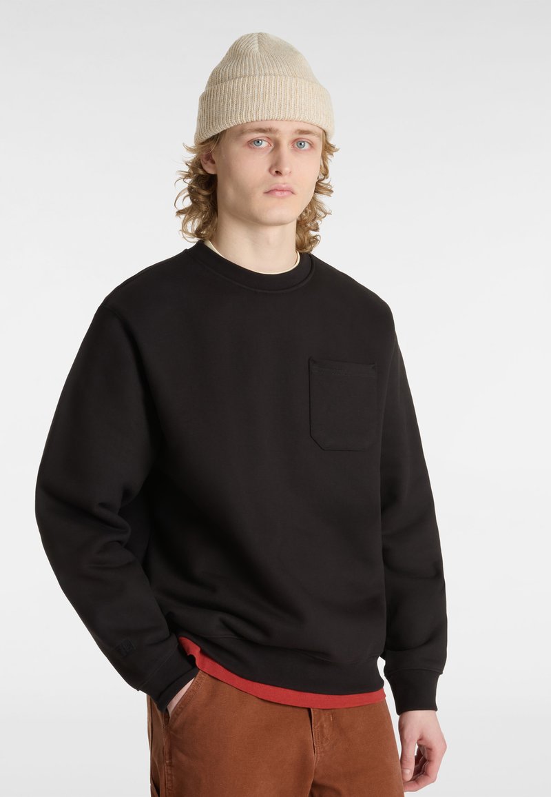 Sweatshirt noir avec un col rond, des poignets côtelés et une poche sur la poitrine gauche. Portez-le sur une chemise de couche rouge, associé à un pantalon marron.